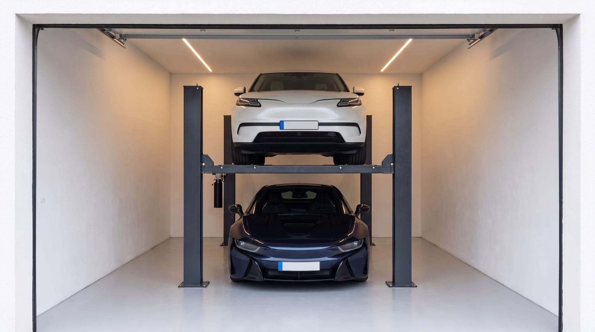 Garage de maison aux murs blancs : doubleur de parking à quatre colonnes anthracite, SUV blanc sur la plateforme levée et coupé bleu nuit garé dessous