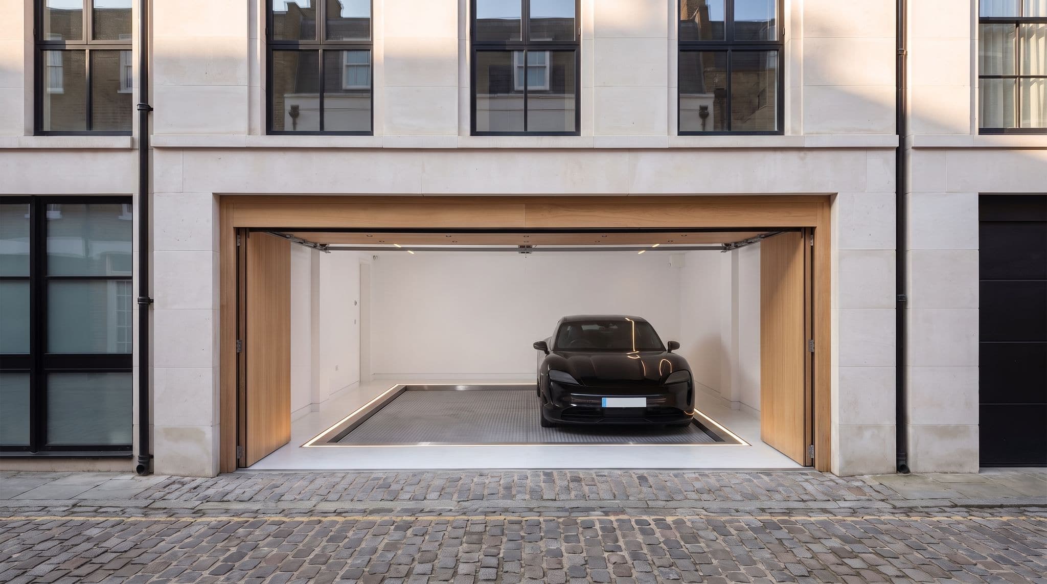 Maison de ville en pierre claire, porte de garage en chêne ouverte : berline noire sur la plateforme d'acier d'un monte-voiture au ras du sol, bordée d'un filet lumineux